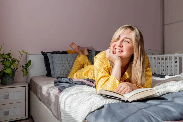 Young woman in yellow pajamas read book while lying on bed in bedroom. Home wellbeing concept. Emotional health of a young woman