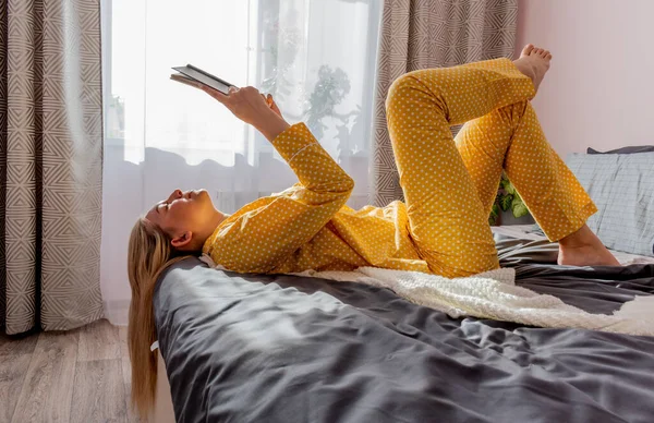 Young woman in yellow pajamas read book while lying on bed in bedroom. Home wellbeing concept. Emotional health of a young woman
