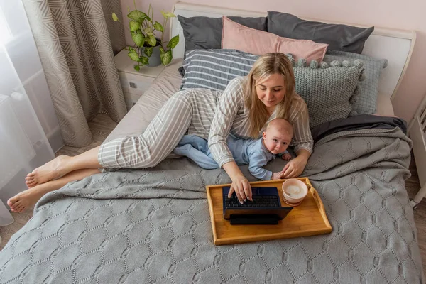 Young woman in comfortable home clothes work at laptop with a baby lying on bed in bedroom. Home wellbeing concept. Emotional health a mom woman