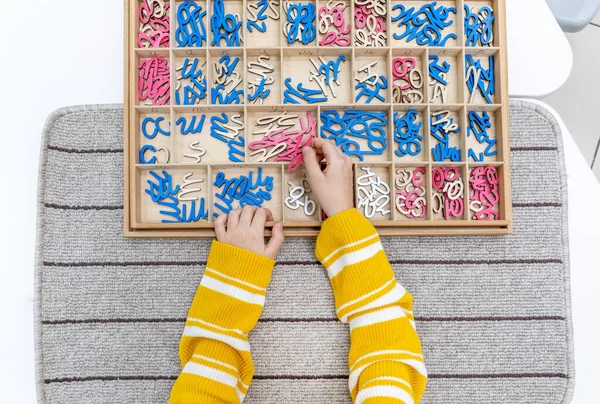 Top view of kids hands building words by using colored Montessori movable alphabet from the wooden tray on gray lined rug. Concept of learning practicing hand coordination and developing motor skills.