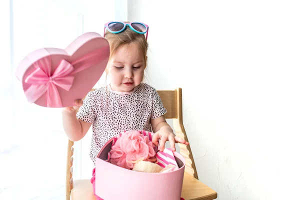 Emotional toddler girl opens a gift. Box in the form of a pink surprise heart. Valentine's Day holiday and gifts concept.
