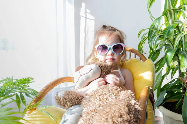 Defoktus. Küçük kız güneşin altında sallanan bir sandalyede oturur, kapalı alandaki bitkilerin arasında. Parlak gölge. Samimi insanlar konsepti. Evde dinlen.