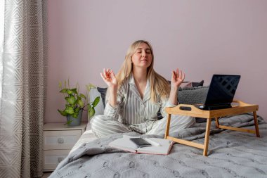 Genç iş kadını, işten sonra internette bilgisayar kullanarak yatakta otururken meditasyon yapıyor. Kız dizüstü bilgisayarın yanında sakin müzik dinliyor. Ev konfor bölgesi ve sağlık konsepti.