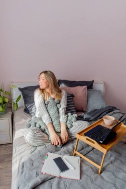 Genç iş kadını, işten sonra internette bilgisayar kullanarak yatakta otururken meditasyon yapıyor. Kız dizüstü bilgisayarın yanında sakin müzik dinliyor. Ev konfor bölgesi ve sağlık konsepti.