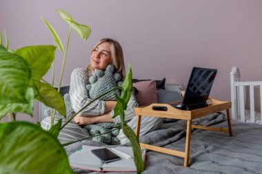 Genç iş kadını, işten sonra internette bilgisayar kullanarak yatakta otururken meditasyon yapıyor. Kız dizüstü bilgisayarın yanında sakin müzik dinliyor. Ev konfor bölgesi ve sağlık konsepti.