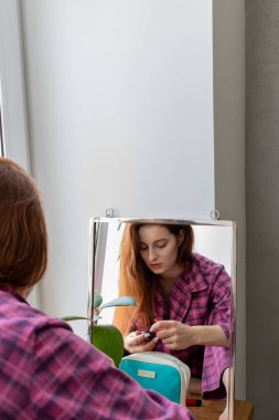 Genç kızıl saçlı kız aynanın önünde pencere eşiğine oturur ve makyaj yapar. Görünüşüne önem verme kavramı. İç mimari