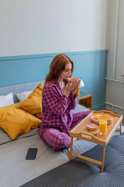 Young woman in pink pajamas has healthy breakfast while lying on bed in bedroom. Home wellbeing concept. Emotional health of a young woman