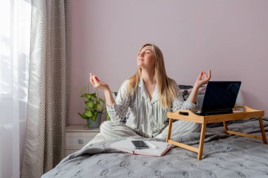 Genç iş kadını, işten sonra internette bilgisayar kullanarak yatakta otururken meditasyon yapıyor. Kız dizüstü bilgisayarın yanında sakin müzik dinliyor. Ev konfor bölgesi ve sağlık konsepti.