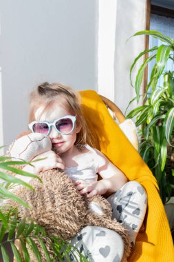 Defoktus. Küçük kız güneşin altında sallanan bir sandalyede oturur, kapalı alandaki bitkilerin arasında. Parlak gölge. Samimi insanlar konsepti. Evde dinlen.