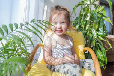 Defoktus. Küçük kız güneşin altında sallanan bir sandalyede oturur, kapalı alandaki bitkilerin arasında. Parlak gölge. Samimi insanlar konsepti. Evde dinlen.