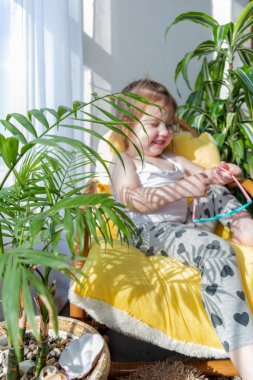 Defoktus. Küçük kız güneşin altında sallanan bir sandalyede oturur, kapalı alandaki bitkilerin arasında. Parlak gölge. Samimi insanlar konsepti. Evde dinlen.