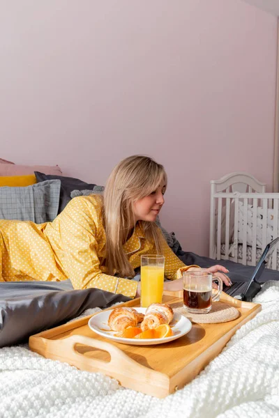 黄色のパジャマの若い女性は ベッドルームでベッドの上に横たわっている間 健康的な朝食と仕事のラップトップを持っています 家庭の幸福の概念 若い女性の感情的な健康 ウェルネス — ストック写真