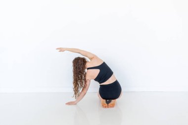 Young diverse attractive woman practicing yoga sitting in Sukhasana pose. Lateral bending exercises. Workout in sportswear. White loft studio background