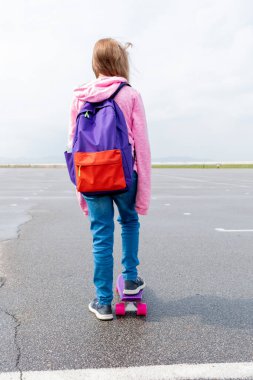 Kot pantolonlu ve pembe kazaklı genç bir kız sokakta kaykaya biniyor. Genç yaşam tarzı konsepti. Uzayı kopyala Arkasında mor bir sırt çantası var..