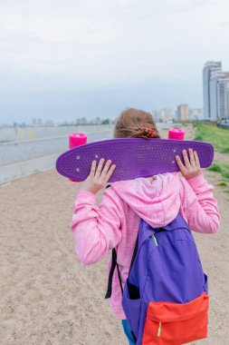 Kot pantolonlu ve pembe kazaklı genç bir kız sokakta bir kaykay tutuyor. Genç yaşam tarzı konsepti. Uzayı kopyala Arkasında mor bir sırt çantası var..