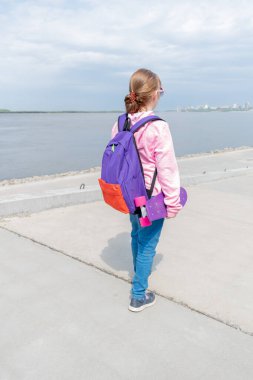 Kot pantolonlu ve pembe kazaklı genç bir kız sokakta bir kaykay tutuyor. Genç yaşam tarzı konsepti. Uzayı kopyala Arkasında mor bir sırt çantası var..