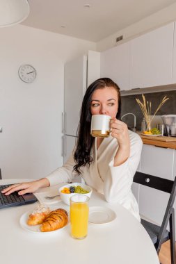 Rahat beyaz cüppeli genç bir kadın mutfaktaki masada otururken dizüstü bilgisayarla çalışıyor. Kahvaltıda evde çalış. İş kadını sosyal mesafeyi koruyor, uzaktan çalışıyor. Yeni normal kavram