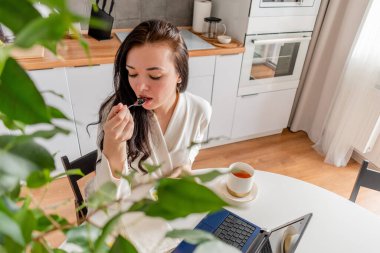 Rahat beyaz cüppeli genç bir kadın mutfaktaki masada otururken dizüstü bilgisayarla çalışıyor. Kahvaltıda evde çalış. İş kadını sosyal mesafeyi koruyor, uzaktan çalışıyor. Yeni normal kavram