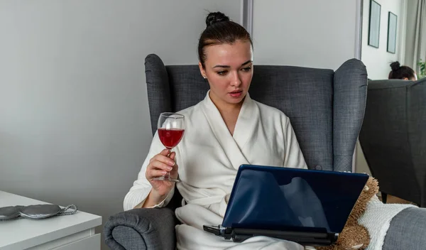 快適な白いローブの若い女性は 彼女の手の中にワインのガラスを保持し ラップトップ上でオンラインの友人と通信します 新常態概念 — ストック写真