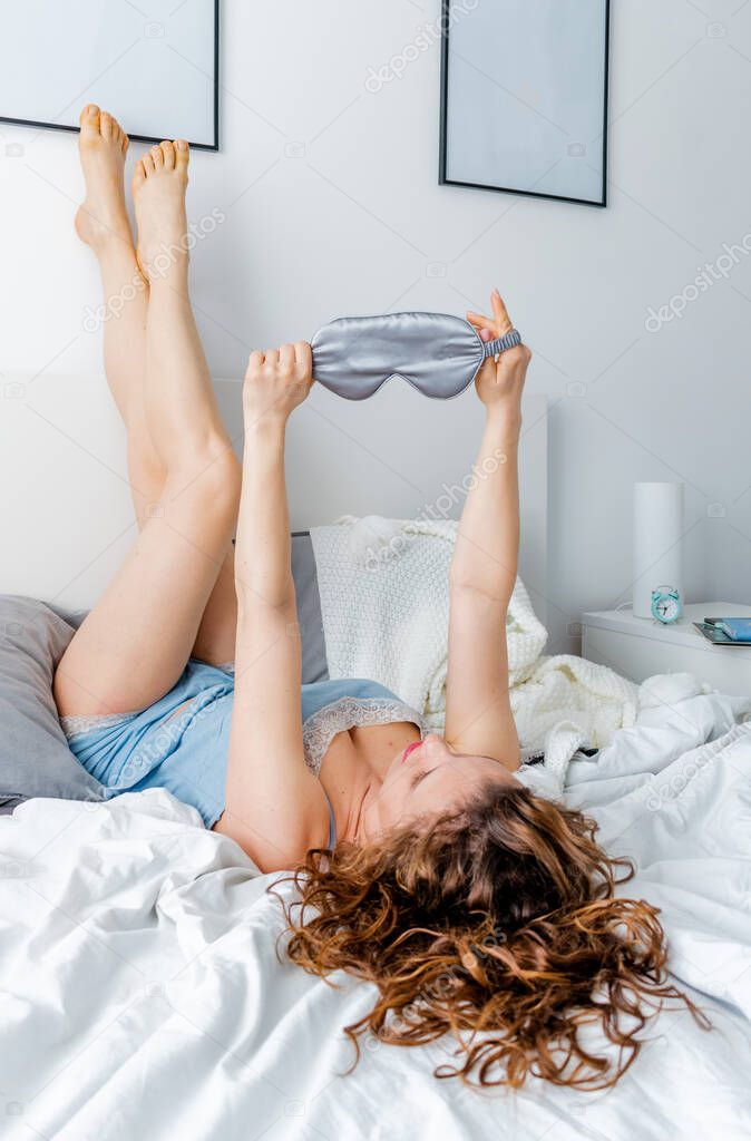 Young ginger hair woman in silk pajamas lies upside down on bed in bedroom. Home wellness concept. Emotional health girl