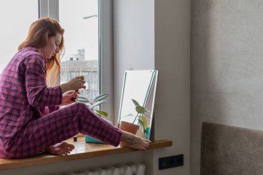 Genç kızıl saçlı kız aynanın önünde pencere eşiğine oturur ve makyaj yapar. Görünüşüne önem verme kavramı. İç mimari