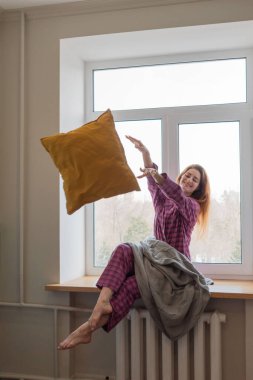 Genç bir kadın rahat pijamalarıyla pencere pervazına oturur ve yastığını fırlatır. Ev hayatı. Yeni bir gerçekliği kabul et.