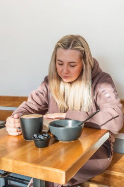 Rahat eşofmanlı genç bir kadın kafede kahve içerken bir kağıt kitap okuyor. Kahvaltıda sağlıklı ve dengeli yemek kavramı. Sosyal ağlardan ve aygıtlardan dinlen.