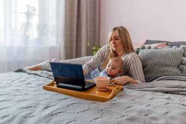 Rahat ev kıyafetleri içinde genç bir kadın laptopta çalışıyor ve yatak odasında bebek yatıyor. Ev refahı kavramı. Duygusal sağlık bir anne kadın