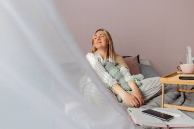 Genç iş kadını, işten sonra internette bilgisayar kullanarak yatakta otururken meditasyon yapıyor. Kız dizüstü bilgisayarın yanında sakin müzik dinliyor. Ev konfor bölgesi ve sağlık konsepti.