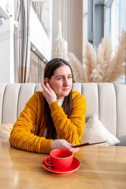 Turuncu renkli hırkalı genç bir kadın, kafenin penceresinin yanında kahve içerken Ebook okuyucusu hakkında elektronik kitap okuyor. Teknoloji ve sanal kütüphane kullanma kavramı. Taşınabilir aygıt.