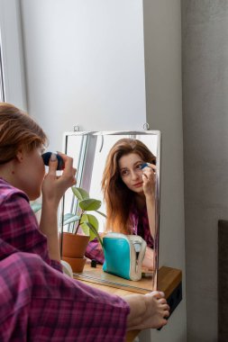 Genç kızıl saçlı kız aynanın önünde pencere eşiğine oturur ve makyaj yapar. Görünüşüne önem verme kavramı. İç mimari