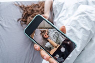 Genç bir kadın uyandı ve pijamalarıyla yatak odasında yatarken selfie çekti. İyi sabahlar. Kız telefon kamerasına bakıyor. Refah kavramı.