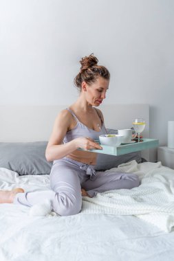 İpek pijamalı genç bir kadın yatakta yatarken sağlıklı kahvaltı yapar. Ev refahı kavramı. Genç bir kadının duygusal sağlığı