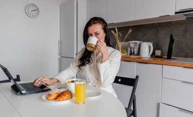 Rahat beyaz cüppeli genç bir kadın mutfaktaki masada otururken dizüstü bilgisayarla çalışıyor. Kahvaltıda evde çalış. İş kadını sosyal mesafeyi koruyor, uzaktan çalışıyor. Yeni normal kavram