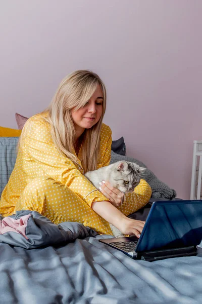 Young woman works on laptop sitting on bed in bedroom next to her pet. Remote work concept. Wellness and mental health concept.