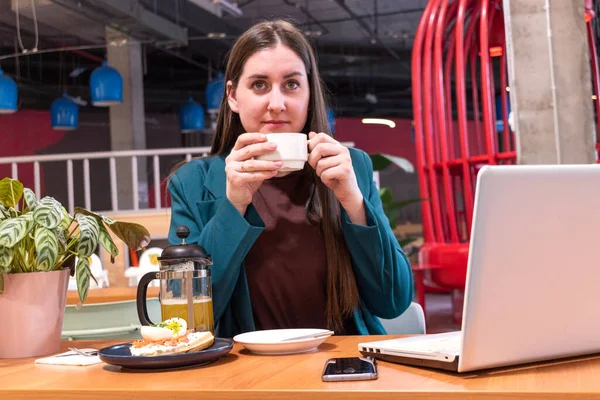 Young woman in suite drinks hot coffee from a mug in a cafe. Businesswoman having light lunch after working hours on laptop.