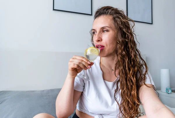 Young woman ginger hair sitting the bed in bedroom and drinks water with lemon. Fresh morning. Wellness and detox concept. Copy space