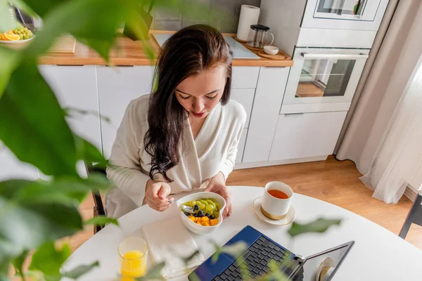 Rahat beyaz cüppeli genç bir kadın mutfaktaki masada otururken dizüstü bilgisayarla çalışıyor. Kahvaltıda evde çalış. İş kadını sosyal mesafeyi koruyor, uzaktan çalışıyor. Yeni normal kavram