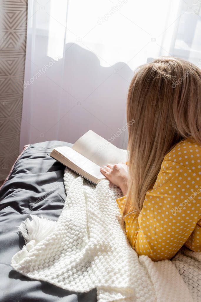 Young woman in yellow pajamas read book while lying on bed in bedroom. Home wellbeing concept. Emotional health of a young woman