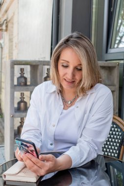 Kafedeki genç kadın bir kafede otururken akıllı telefonla çalışıyor. Beyaz elbiseli sarışın iş kadını. Yeni normal kavram