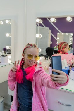 Genç kız kameraya bakıp telefonuna bakarak selfie çekiyor. Çocuk kuaförde saçını yaptıktan sonra kendi fotoğrafını çekiyor. Saçları örgülü.