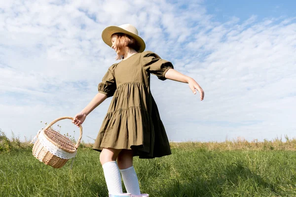 Girl in a linen dress and a straw hat is dancing on a flower field. Wicker basket in hands. Wellness and freedom concept. Teenager in nature