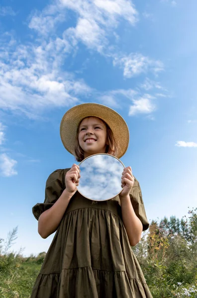 Girl is holding a mirror in which the sky is reflected. Glade with flowers. Wellness and freedom concept. Teenager in nature