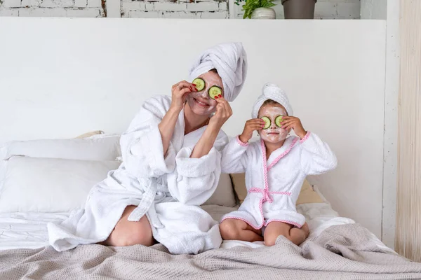 母親と娘は化粧品マスクとキュウリでベッドに座っています 服は白いテリーローブです 家族の看護コミュニケーション ウェルネスコンセプト — ストック写真