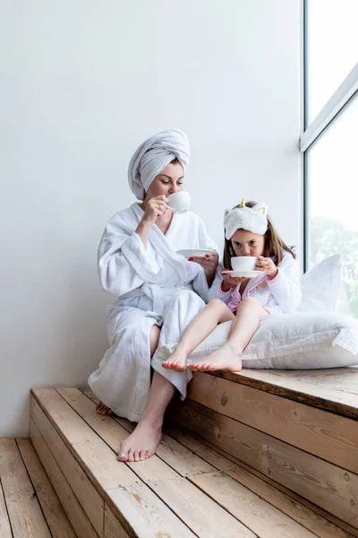 母と娘は窓のそばに座り 白い衣と白いタオルを頭にかぶってお茶を飲んでいます ウェルネスの概念 家で過ごした時間 — ストック写真