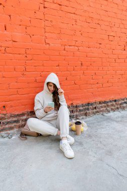 Farklılık gösteren yetişkin bir kadın, sokakta oturur ve termo bardaktan kahve içer, telefon kamerasına bakar ve selfie çeker. Sıfır kayıp. Modern teknolojileri kullanma kavramı.