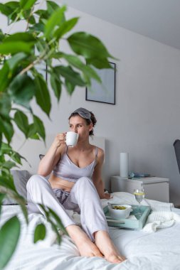 İpek pijamalı genç bir kadın yatakta yatarken sağlıklı kahvaltı yapar. Ev refahı kavramı. Genç bir kadının duygusal sağlığı