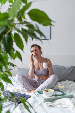 Rahat leylak pijamalı genç bir kadın bilgisayarda çalışıyor ve yatak odasında yatarken cep telefonuyla konuşuyor. İş kadını sosyal mesafeyi koruyor ve uzaktan çalışıyor. Yeni normal kavram