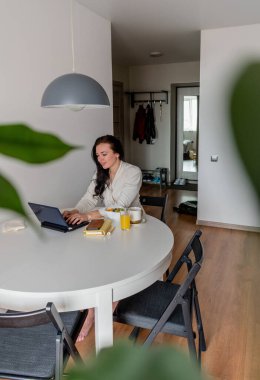 Rahat beyaz cüppeli genç bir kadın mutfaktaki masada otururken dizüstü bilgisayarla çalışıyor. Kahvaltıda evde çalış. İş kadını sosyal mesafeyi koruyor, uzaktan çalışıyor. Yeni normal kavram