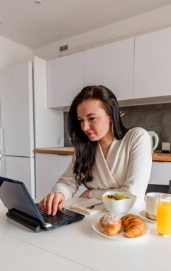 Rahat beyaz cüppeli genç bir kadın mutfaktaki masada otururken dizüstü bilgisayarla çalışıyor. Kahvaltıda evde çalış. İş kadını sosyal mesafeyi koruyor, uzaktan çalışıyor. Yeni normal kavram